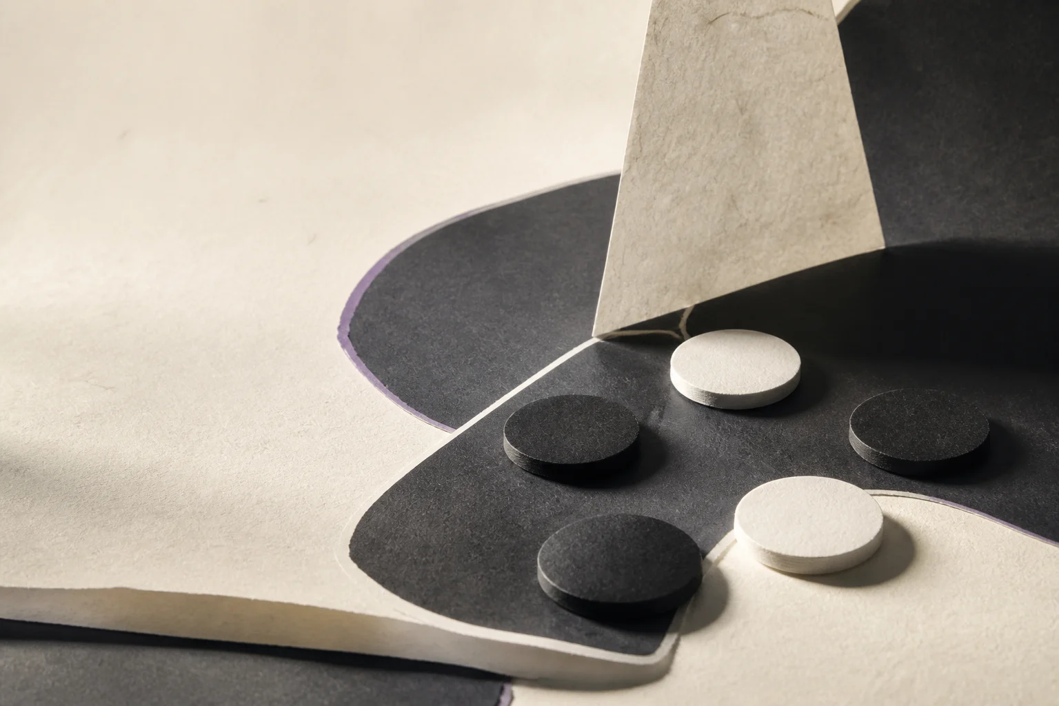 Black and white tactile Go stones arranged across folded cream paper and charcoal fields, divided by a fine white line.